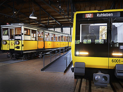 Blick auf zwei nebeneinander stehende U-Bahnwagen. Beide sind gelb. Der Wagen rechts sieht sehr modern aus, der Wagen links ist ein historisches Fahrzeug.