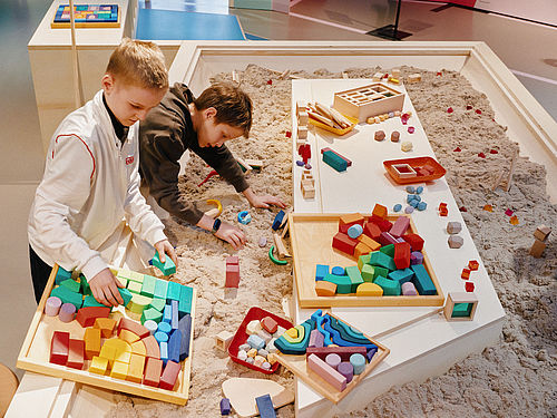 In einem großen Sandkasten liegen viele bunte Bauklötze. Zwei Jungen bauen mit dem Sand und den Bauelementen eine kleine Stadt.