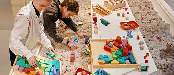 In einem großen Sandkasten liegen viele bunte Bauklötze. Zwei Jungen bauen mit dem Sand und den Bauelementen eine kleine Stadt.
