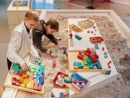 In einem großen Sandkasten liegen viele bunte Bauklötze. Zwei Jungen bauen mit dem Sand und den Bauelementen eine kleine Stadt.