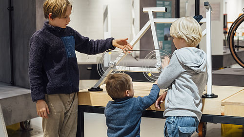 Drei Kinder stehen in einem Ausstellungsraum vor einem schematisch vereinfachten Nachbau eines Fahrrads. Ein Junge berührt die dort dargestellte Fahrradkette.