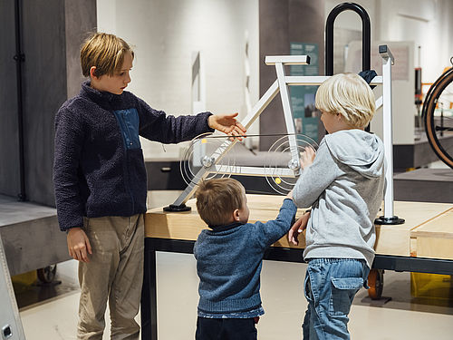Three children stand in an exhibition room in front of a schematically simplified replica of a bicycle. One boy touches the bicycle chain depicted there.