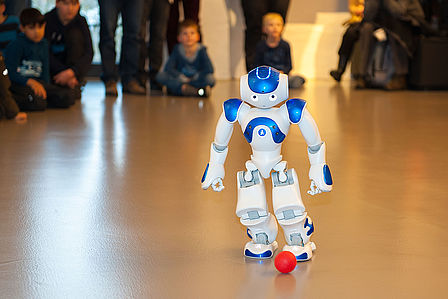 Der weiß-blaue Roboter NAO spielt mit einem kleinen roten Ball. Der etwa 60 Zentimeter große humanoide Roboter geht auf den Ball zu und versucht ihn zu treten. Im Hintergrund verfolgen Kinder und Erwachsene die Vorführung.