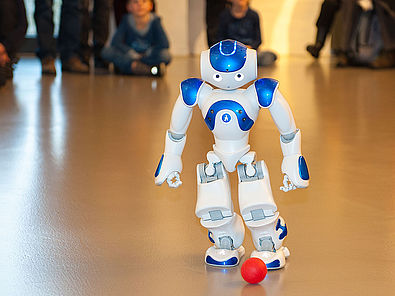 Der weiß-blaue Roboter NAO spielt mit einem kleinen roten Ball. Der etwa 60 Zentimeter große humanoide Roboter geht auf den Ball zu und versucht ihn zu treten. Im Hintergrund verfolgen Kinder und Erwachsene die Vorführung.