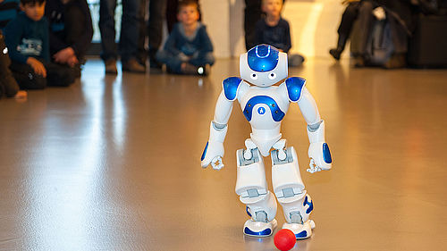 Der weiß-blaue Roboter NAO spielt mit einem kleinen roten Ball. Der etwa 60 Zentimeter große humanoide Roboter geht auf den Ball zu und versucht ihn zu treten. Im Hintergrund verfolgen Kinder und Erwachsene die Vorführung.