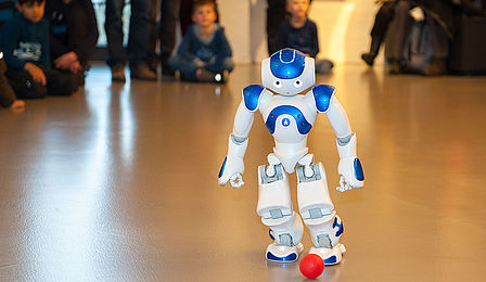 Der weiß-blaue Roboter NAO spielt mit einem kleinen roten Ball. Der etwa 60 Zentimeter große humanoide Roboter geht auf den Ball zu und versucht ihn zu treten. Im Hintergrund verfolgen Kinder und Erwachsene die Vorführung.
