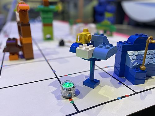 Ein kleiner blauleuchtenden Roboter fährt über einen Linennetz auf dem kleine Gebäude aus Steckbausteinen stehen.