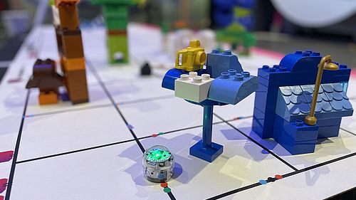Ein kleiner blauleuchtenden Roboter fährt über einen Linennetz auf dem kleine Gebäude aus Steckbausteinen stehen.