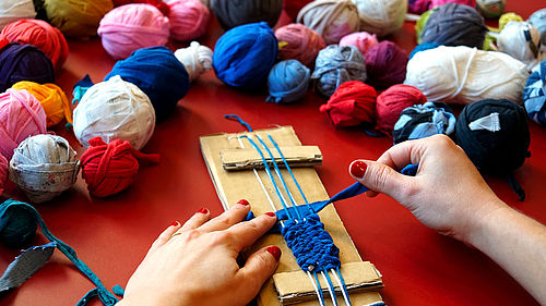 Eine Person sitzt an einem Tisch und webt an einem selbstgebauten Webrahmen aus Pappe aus einem breiten, blauen Stück Stoff ein Band. Von der Person sind nur die Hände zu sehen. Auf dem Tisch liegen viele bunte Knäule aus Stoffresten.