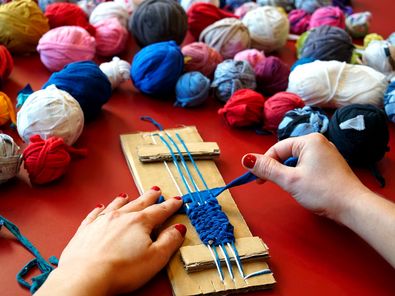 Eine Person sitzt an einem Tisch und webt an einem selbstgebauten Webrahmen aus Pappe aus einem breiten, blauen Stück Stoff ein Band. Von der Person sind nur die Hände zu sehen. Auf dem Tisch liegen viele bunte Knäule aus Stoffresten.