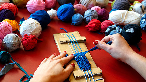 Eine Person sitzt an einem Tisch und webt an einem selbstgebauten Webrahmen aus Pappe aus einem breiten, blauen Stück Stoff ein Band. Von der Person sind nur die Hände zu sehen. Auf dem Tisch liegen viele bunte Knäule aus Stoffresten.