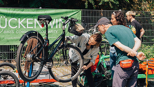 Mehrere Menschen gruppieren sich im Freien um ein Fahrrad, das an einem Gestell hängt. Eine Frau erklärt etwas zur Funktionsweise des Rads.
