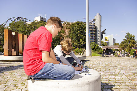 Zwei Jungen sitzen auf dem Vorplatz des Deutschen Technikmuseums und beugen sich über ihre Notizen. Im Hintergrund ist das Rotorblatt zu sehen.