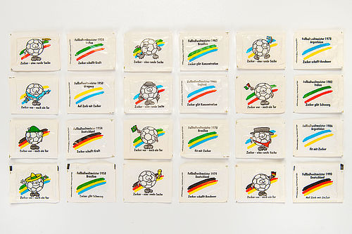 24 weiße rechteckige Zuckertütchen sind chronologisch in der Reihenfolge ihres Fußballweltmeister-Titels sortiert. Sie sind bedruckt mit Motiven wie Fußbällen, die landestypische Hüte tragen oder Fahnen schwenken sowie den jeweiligen Nationalfarben der jeweiligen Weltmeister und Werbesprüchen wie „Zucker – eine runde Sache“ oder „Zucker schafft Ausdauer“.