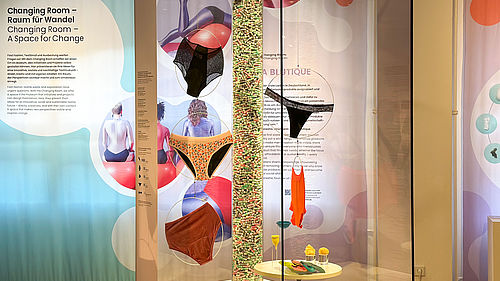 Blick auf die Vitrine im Ausstellungsbereich "Changing Room": Zu sehen sind verschiedene Modelle von Perioden-Unterwäsche, Textil-Binden, Menstruations-Cups und Tampons. Im Hintergrund ist der Schriftzug "Changing Room" zu lesen.