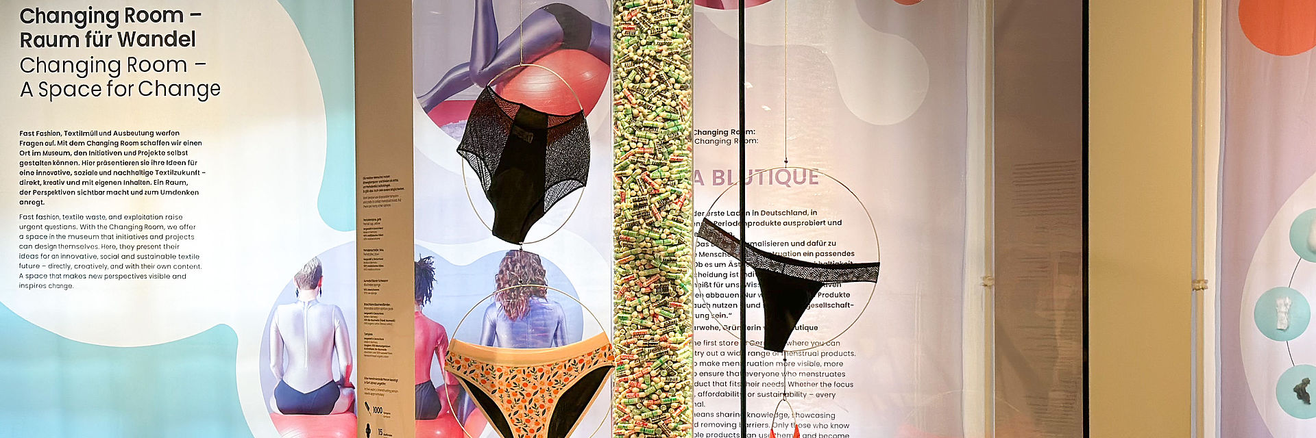Blick auf die Vitrine im Ausstellungsbereich "Changing Room": Zu sehen sind verschiedene Modelle von Perioden-Unterwäsche, Textil-Binden, Menstruations-Cups und Tampons. Im Hintergrund ist der Schriftzug "Changing Room" zu lesen.