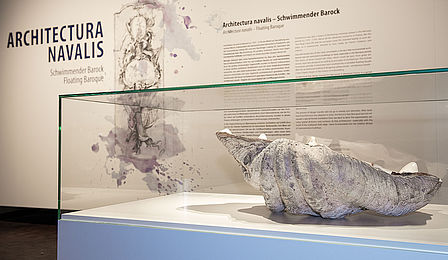 Blick in die Sonderausstellung: Im Vordergrund eine Vitrine mit einer Riesenmuschel, im Hintergrund eine Texttafel mit der Zeichnung eines barocken Schiffshecks und dem Titel „Architectura navalis“.