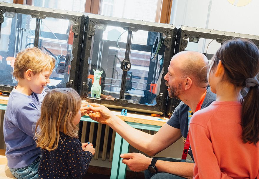 Ein Mann und drei Kinder sitzen vor einer Reihe von 3D-Druckern. Der Mann erklärt etwas, die Kinder haben Spaß und hören zu.