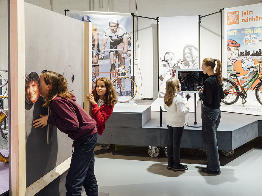 Several people in an exhibition space. In the foreground, two young girls standing around a wooden wall with holes in it; in the background, a young woman and a little girl talking in front of an exhibition unit with bicycles and pictures of cyclists.