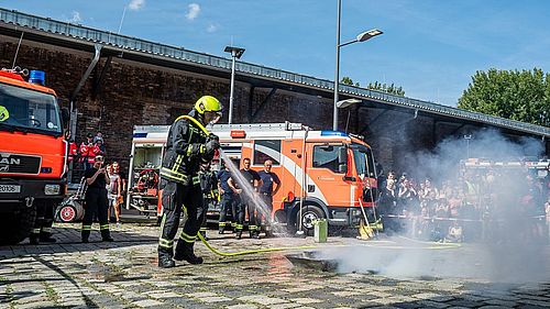 Ein Feuerwehrmann löscht mit einem starken Wasserstrahl ein kleines Demonstrationsfeuer in der Ladestraße des Deutschen Technikmuseums. Im Hintergrund stehen Rettungsfahrzeuge und Besucherinnen und Besucher des Blaulichtfestes.