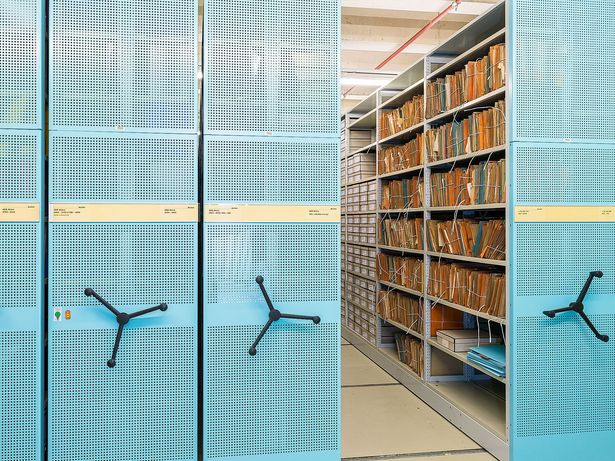 Modernes Roll-Regalsystem aus Metall, die Außenseiten sind Hellblau. Der rechte Regalteil ist aufgeschoben, so dass man im Regal zusammengeschnürte Akten und graue Archivboxen sehen kann.