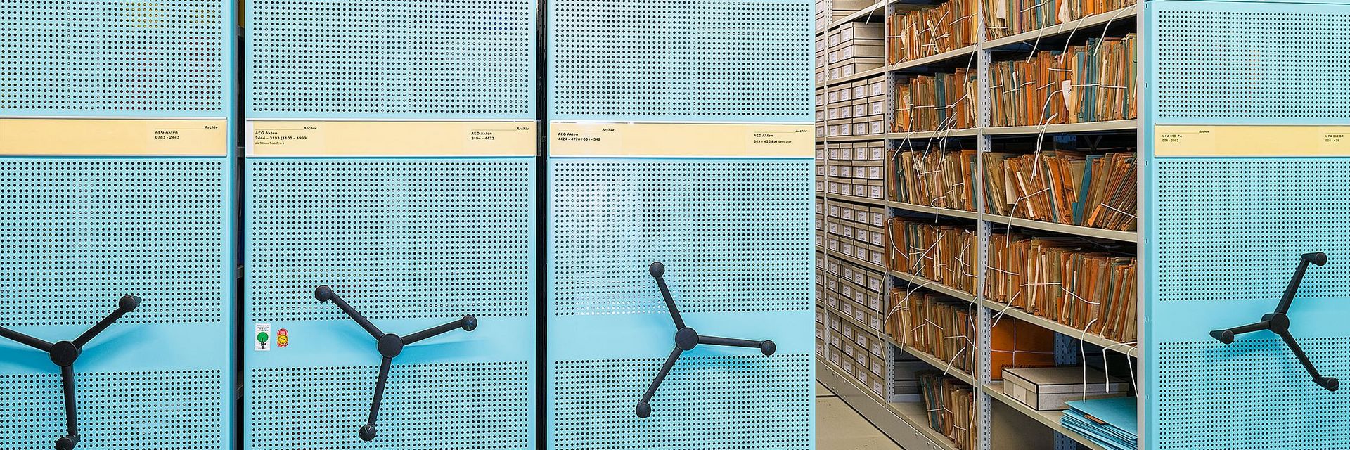 Modernes Roll-Regalsystem aus Metall, die Außenseiten sind Hellblau. Der rechte Regalteil ist aufgeschoben, so dass man im Regal zusammengeschnürte Akten und graue Archivboxen sehen kann.