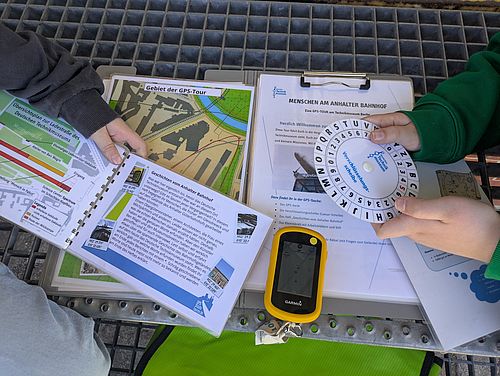 Man sieht die Materialien für die GPS-Tour: Ein GPS-Gerät, eine Verschlüsselungsscheibe und zwei Informationshefte.