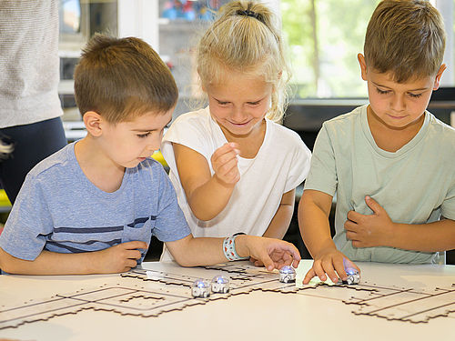 Drei Kinder sitzen an einem Tisch und beschäftigen sich mit dem kleinen Roboter Ozobot, der seine Anweisungen über Sensoren erhält.