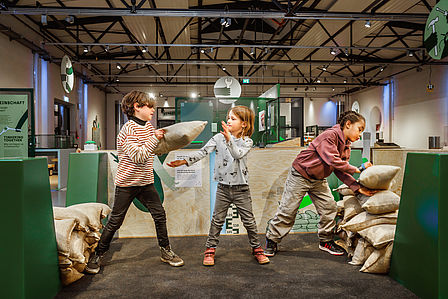 Drei Kinder reparieren in einer Ausstellungshalle einen Spielzeug-Deich aus Sandsäcken. Sie bilden eine Kette und reichen sich die Säcke, um eine Lücke im Deich zu flicken.