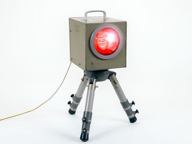 A rectangular, elongated metal flash unit is mounted on a tripod. The legs are painted grey while the housing is somewhat darker and melds into an olive green color. There is a red round glass disk on the front of the flash unit.