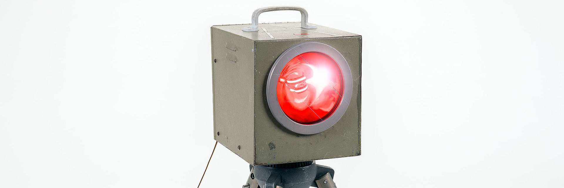A rectangular, elongated metal flash unit is mounted on a tripod. The legs are painted grey while the housing is somewhat darker and melds into an olive green color. There is a red round glass disk on the front of the flash unit.