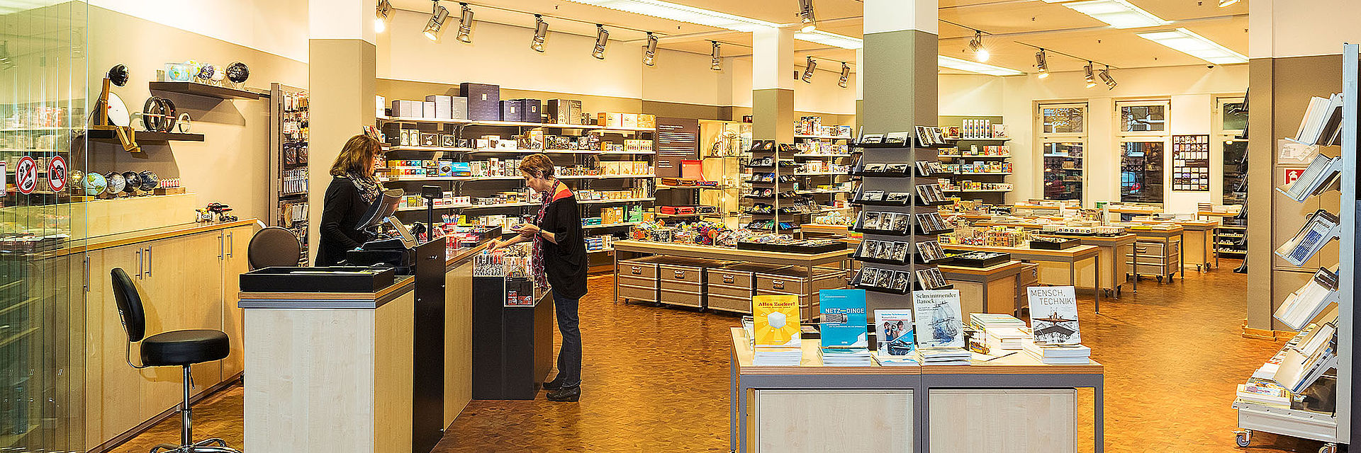 Blick in einen hellen, freundlichen Shop mit Souvenirs und Büchern. Links ist ein Kassentresen, an dem eine Verkäuferin und eine Kundin stehen.