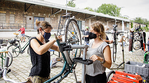 Zwei Frauen schrauben an einem Fahrrad. Im Hintergund arbeiten weitere Personen an Fahrrädern.