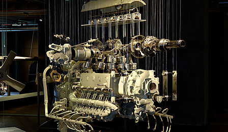 Vor einem dunklen Hintergrund hängen die beleuchteten Einzelteile eines Motors an dünnen Fäden.