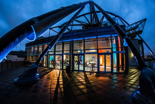 Die Terrasse, die zur Eventfläche AUSGUCK gehört ist regennass und liegt im Dunkeln. Im letzten Tageslicht erscheinen die spinnenartigen Architekturelemente als Schattenriss. Der Blick fällt mittig, durch die Glasfassade, auf den farbig beleuchteten Innenraum.