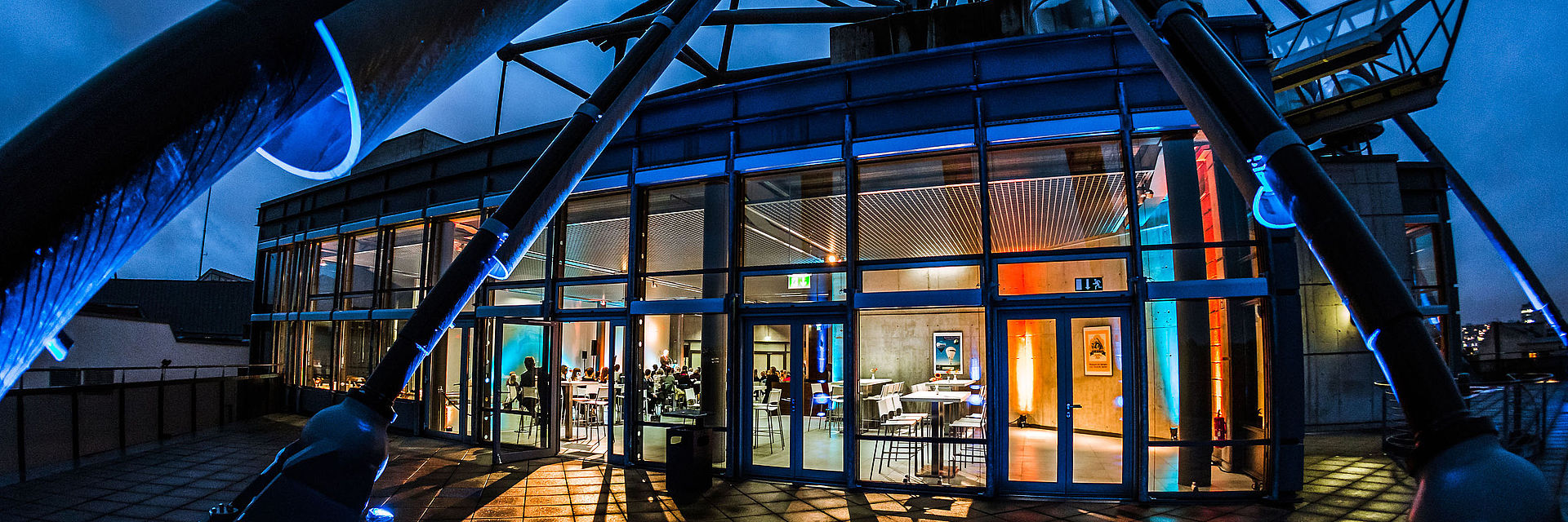 Die Terrasse, die zur Eventfläche AUSGUCK gehört ist regennass und liegt im Dunkeln. Im letzten Tageslicht erscheinen die spinnenartigen Architekturelemente als Schattenriss. Der Blick fällt mittig, durch die Glasfassade, auf den farbig beleuchteten Innenraum.