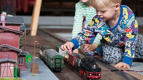 Zwei Kinder sitzen neben einer Spur-0-Modelleisenbahn auf einem Teppich. Auf den Gleisen stehen eine schwarz-rote Lokomotive und mehrere Anhänger. Eines der Kinder belädt einen offenen Anhänger mit kleinen, buntverpackten Süßigkeiten.