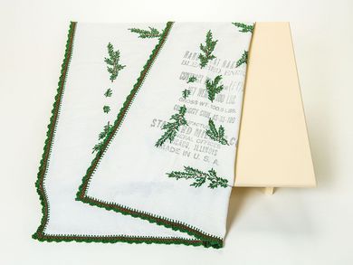 Die quadratische Tischdecke besteht aus weißem Baumwollstoff. Sie ist mit grünen Tannenzweigen bestickt, die in Handarbeit aufgebracht wurden. Auffällig ist ein englischsprachiger Aufdruck auf einem Teil des Tischtuchs. Er zeigt, dass die Tischdecke zuvor ein Mehlsack war.