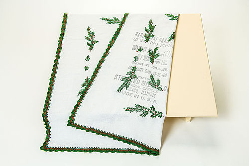 Die quadratische Tischdecke besteht aus weißem Baumwollstoff. Sie ist mit grünen Tannenzweigen bestickt, die in Handarbeit aufgebracht wurden. Auffällig ist ein englischsprachiger Aufdruck auf einem Teil des Tischtuchs. Er zeigt, dass die Tischdecke zuvor ein Mehlsack war.