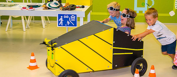 Drei Kinder fahren eine gelb-schwarze Seifenkiste. Das Fahrzeug besteht aus vorgefertigten Holzplatten mit vier Reifen. Auf die Front wurde ein lächelndes Gesicht gemalt. Ein Mädchen sitzt mit einer getönten Brille im Gefährt und steuert es, während zwei Jungen die Kiste anschieben.