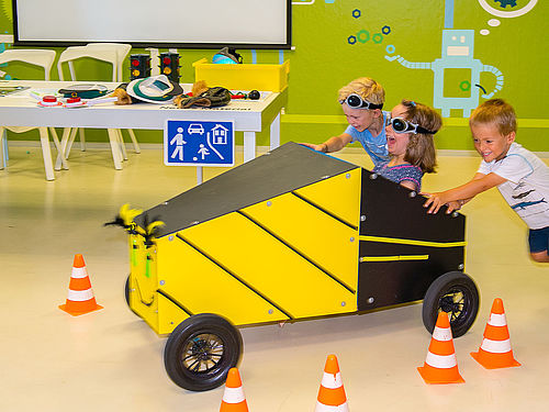 Drei Kinder fahren eine gelb-schwarze Seifenkiste. Das Fahrzeug besteht aus vorgefertigten Holzplatten mit vier Reifen. Auf die Front wurde ein lächelndes Gesicht gemalt. Ein Mädchen sitzt mit einer getönten Brille im Gefährt und steuert es, während zwei Jungen die Kiste anschieben.