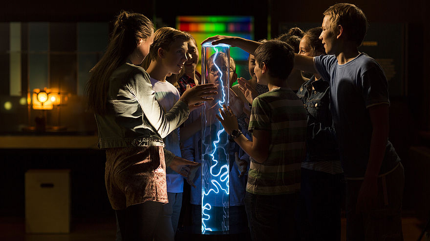 Eine Gruppe von Kindern steht in einem dunklen Raum um eine transparente Säule herum. Im Inneren der Säule leuchtet entlang der gesamten Höhe ein blauer Blitz. Einige Kinder berühren die Plasmasäule mit ihren Händen.