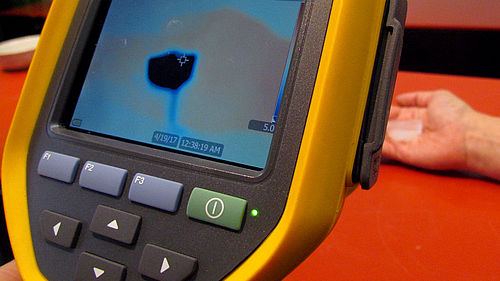 Hinter einem Messgerät mit Display und acht Tasten ist unscharf eine Hand zu erkennen. In dieser liegt ein kleiner durchsichtiger eckiger Gegenstand. Das Display zeigt ein Bild in Rot- und Blautönen, mittig ist ein blauer Würfel erkennbar. 