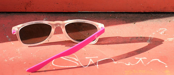Eine zerbrochene Sonnenbrille mit pinkem Bügel liegt auf dem Boden. Dahinter eine weiß-orangene Wand. Es gibt mehrere Dreiecks-Formen in dem Foto.m Fotos 
