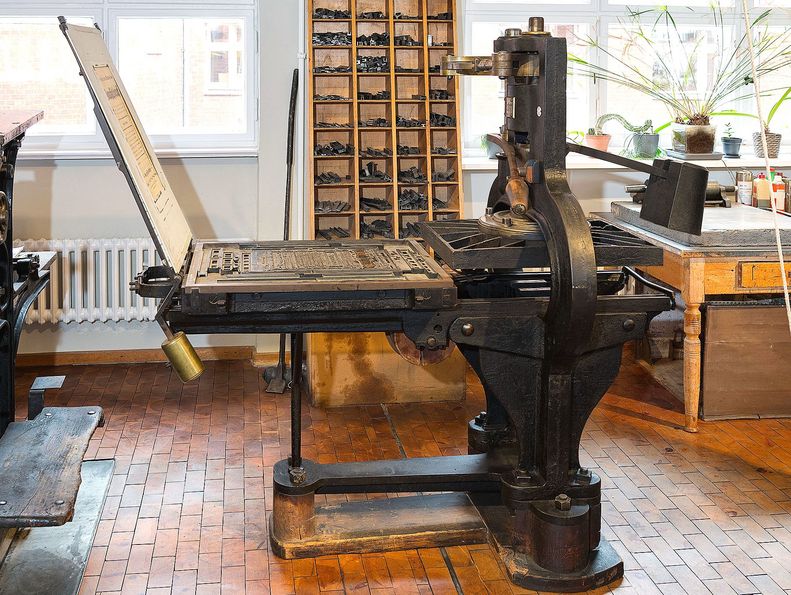 Rechts ist die mit einem Handhebel betriebene gusseiserne Presse zu sehen; links die aufgeklappte Druckplatte mit den Metallbuchstaben.