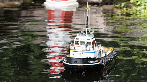 Ein kleines Schiffsmodel fährt auf einem ruhigen Gewässer, im Hintergrund sieht man das Modell eines Leuchtturms, das sich im Wasser spiegelt.