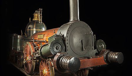 Eine mächtige Dampflokomotive aus metallenen und hölzernen Bauteilen vor schwarzem Hintergrund