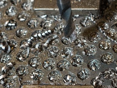 Der Metallbohrer hat zahlreiche Abdrücke in der Platte des Werkzeugtisches hinterlassen. Auf dem Tisch liegen Metallspiralen und kleine Bruchstücke als Abfallprodukte des Bohrvorgangs.