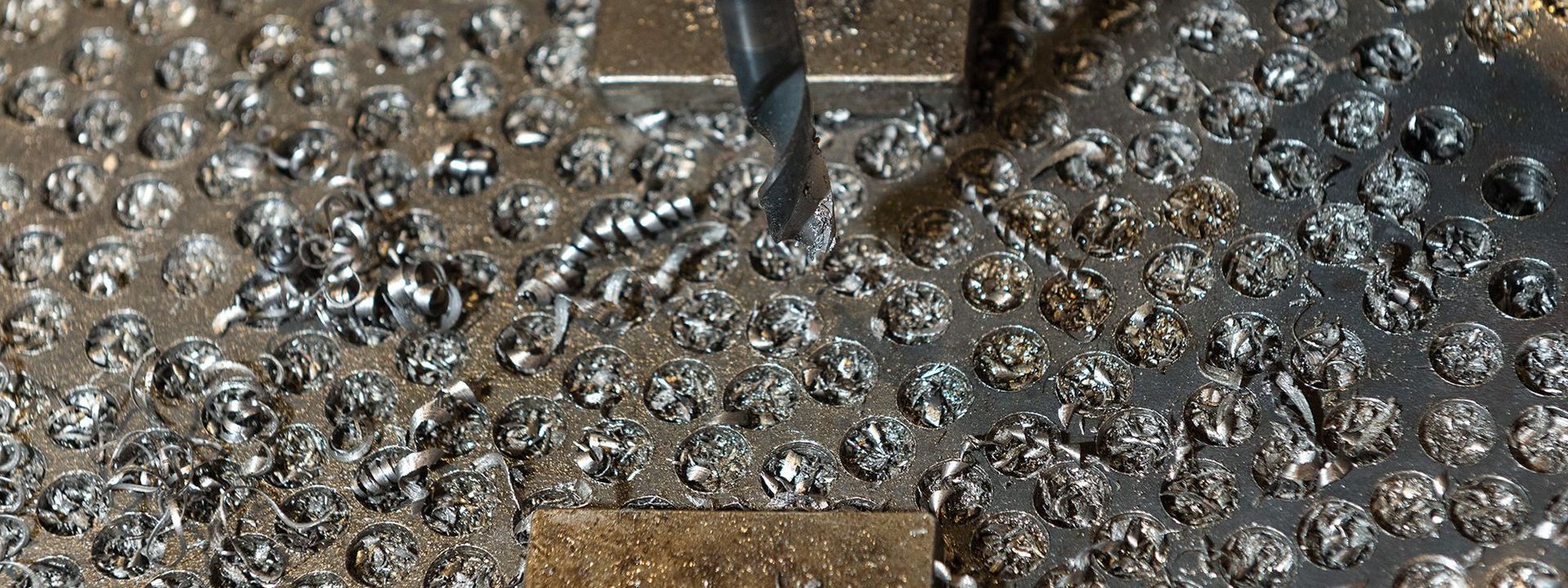 Der Metallbohrer hat zahlreiche Abdrücke in der Platte des Werkzeugtisches hinterlassen. Auf dem Tisch liegen Metallspiralen und kleine Bruchstücke als Abfallprodukte des Bohrvorgangs.