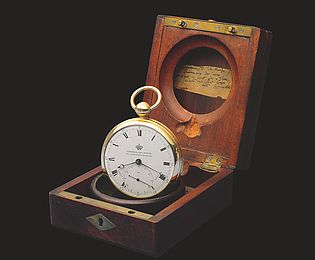 Der Taschenchronometer steht aufrecht in seinem hölzernen Kästchen vor schwarzem Hintergrund.